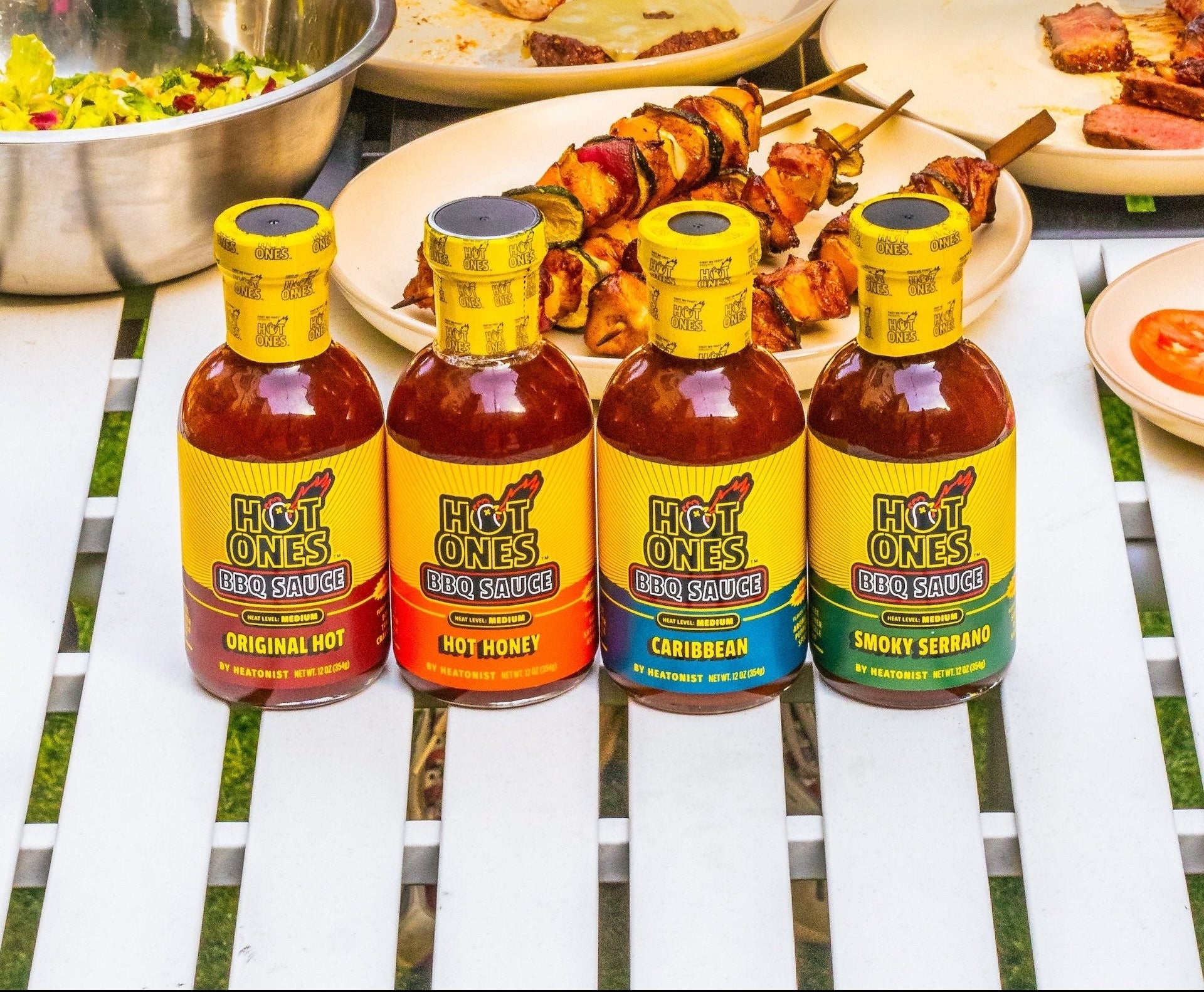 Four bottles of Hot Ones BBQ Sauce lined up on a white slatted table, surrounded by grilled meats, burgers, and other barbecue dishes in an outdoor dining setting.